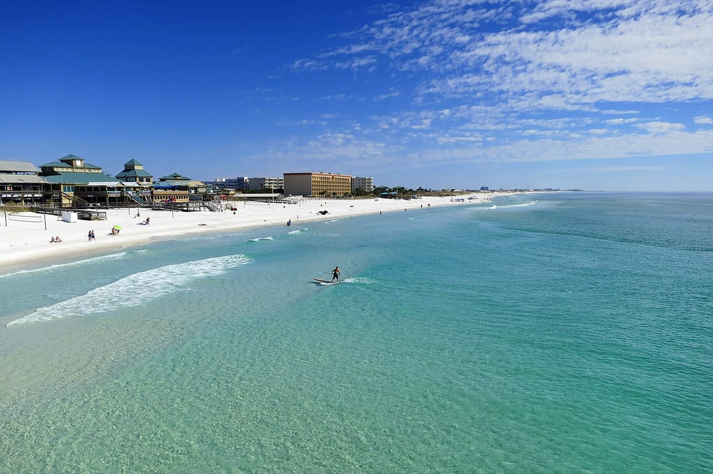 A beach scene with turquoise waters and a white sandy shore. A person is paddleboarding in the clear water. There are buildings along the coast and a partly cloudy blue sky overhead.