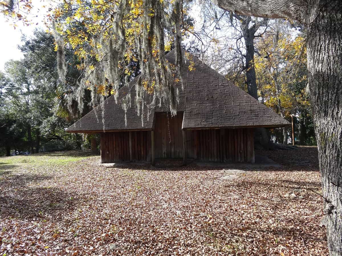 A historical wooden structure with a steeply pitched roof stands amid a leafy, forested area. The ground is covered with autumn leaves, and Spanish moss hangs from the trees, creating a rustic and serene atmosphere.