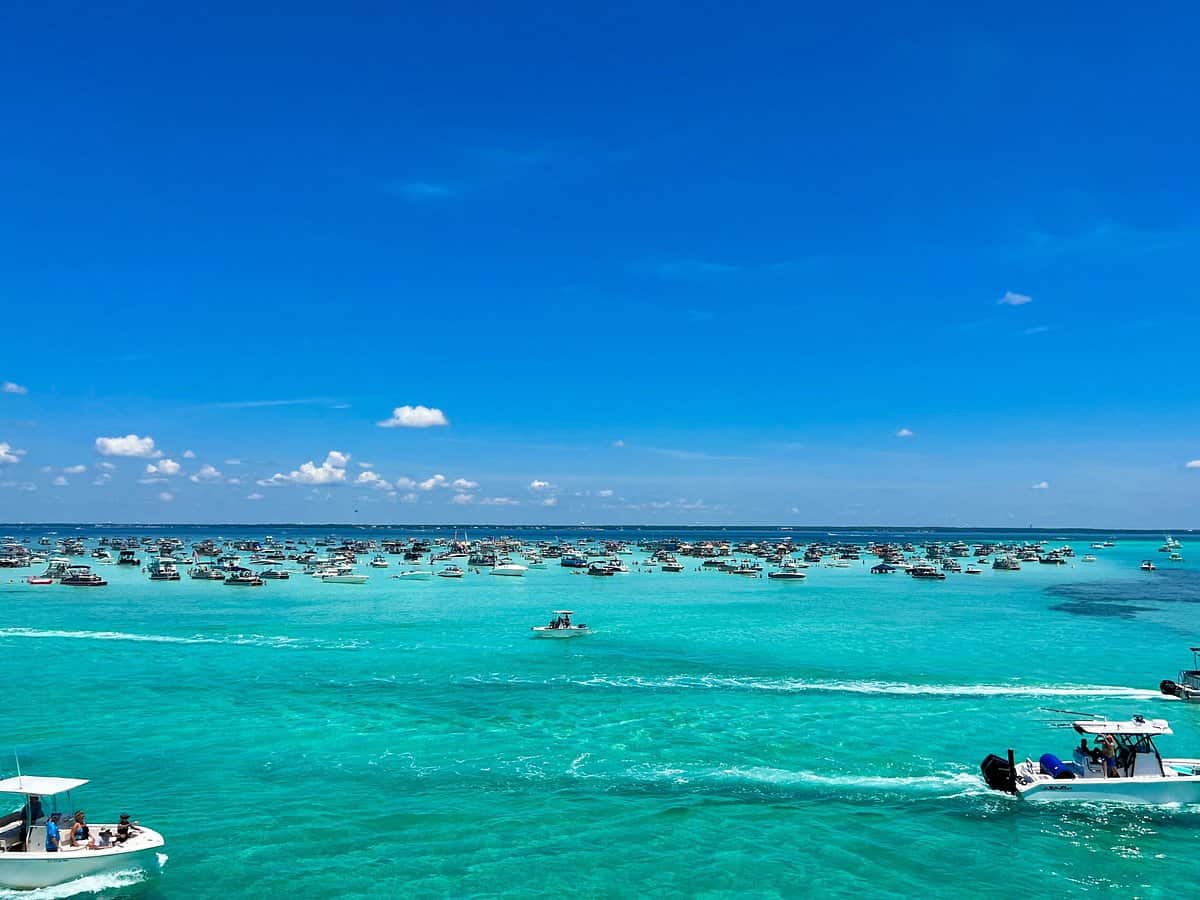 A vibrant blue ocean with numerous small boats scattered across the water beneath a clear blue sky. The sea is calm, reflecting the hue of the sky, and the boats create gentle ripples on the surface.