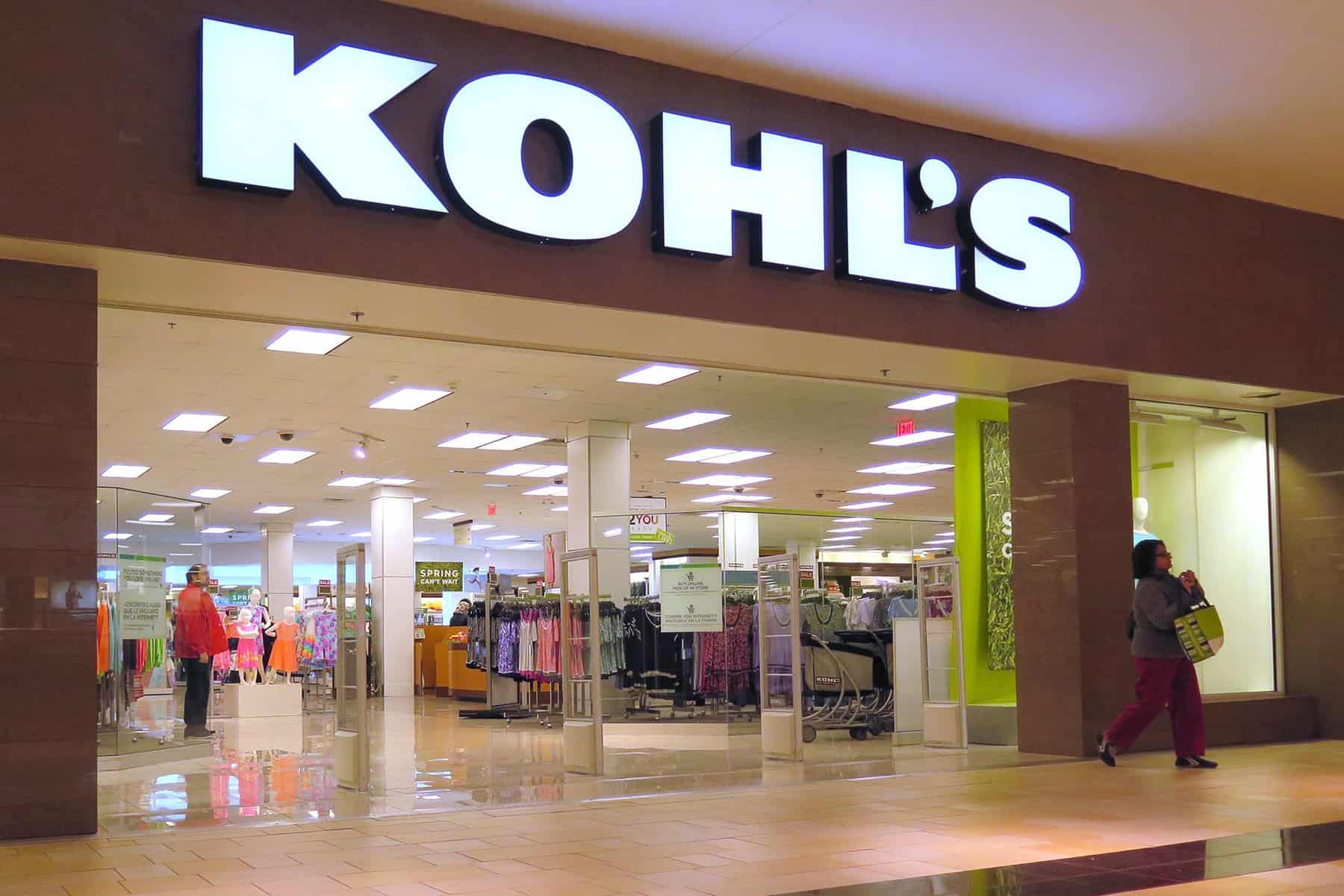 Storefront of Kohl's in a mall. The entrance is wide with glass doors and the store logo prominently displayed above. Inside, there are clothes racks visible, and a person is walking by carrying a shopping bag.