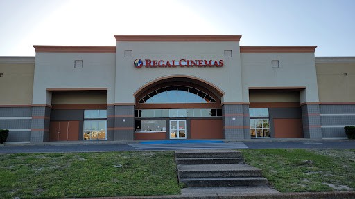 A Regal Cinemas building with a large entrance, featuring multiple doors and a prominent arched window. The facade is a mix of beige and gray with red accents. The foreground includes a small grassy area and concrete steps.