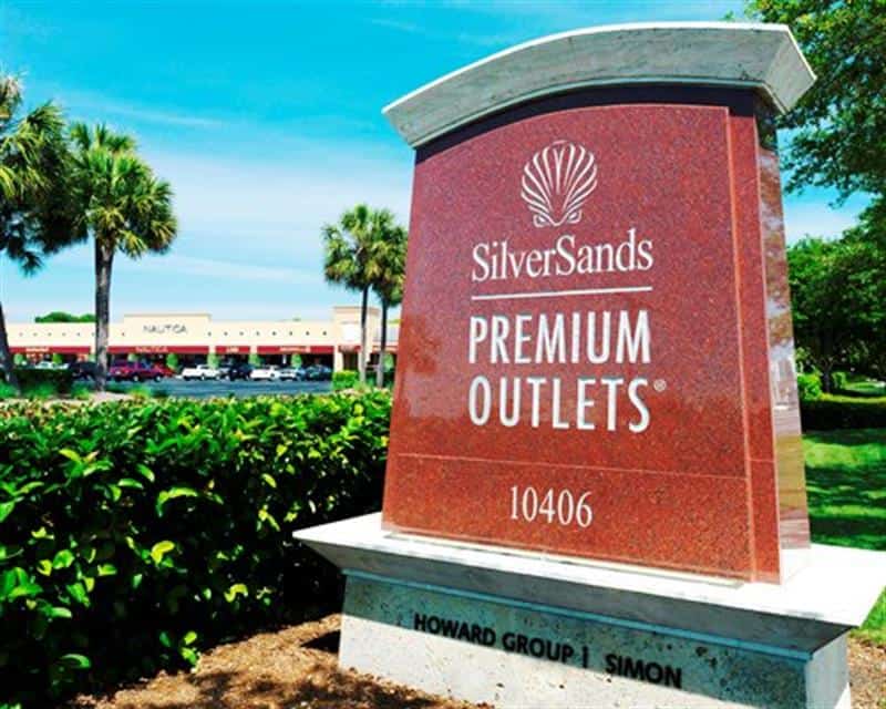 Sign for Silver Sands Premium Outlets surrounded by green bushes, with trees and a shopping area visible in the background under a clear blue sky.