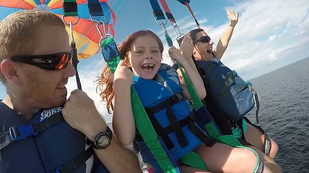 A young girl, a man, and a woman are tandem parasailing over the ocean. They wear harnesses and life vests attached to a colorful parachute. The girl is smiling widely, and the woman waves her hand excitedly.
