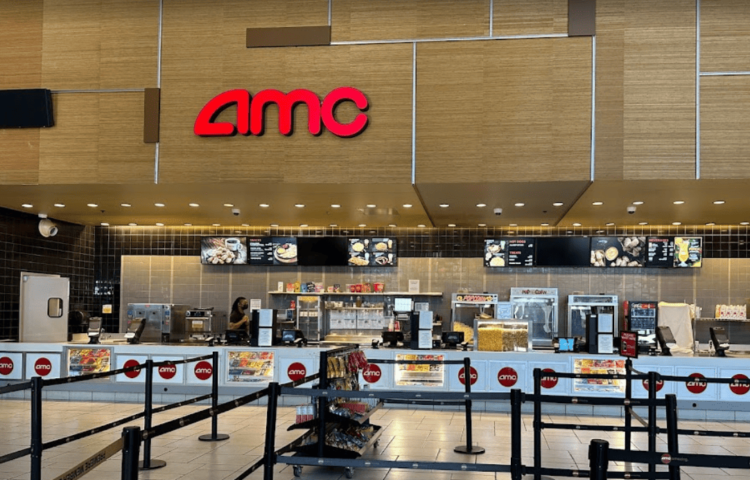 Inside a movie theater concession stand with a large AMC logo on a wooden wall. The counter displays various snacks, popcorn, and drinks. Digital menu screens are above, and queue barriers guide customers.