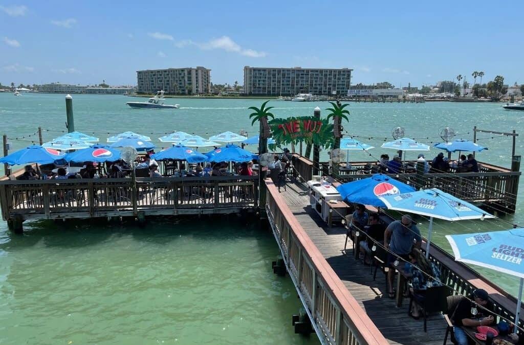 Outdoor dining area on wooden docks overlooking a calm body of water. Patrons sit under blue umbrellas with logos. A boat is passing by in the distance. Several buildings are visible across the water, and palm trees flank a sign reading 
