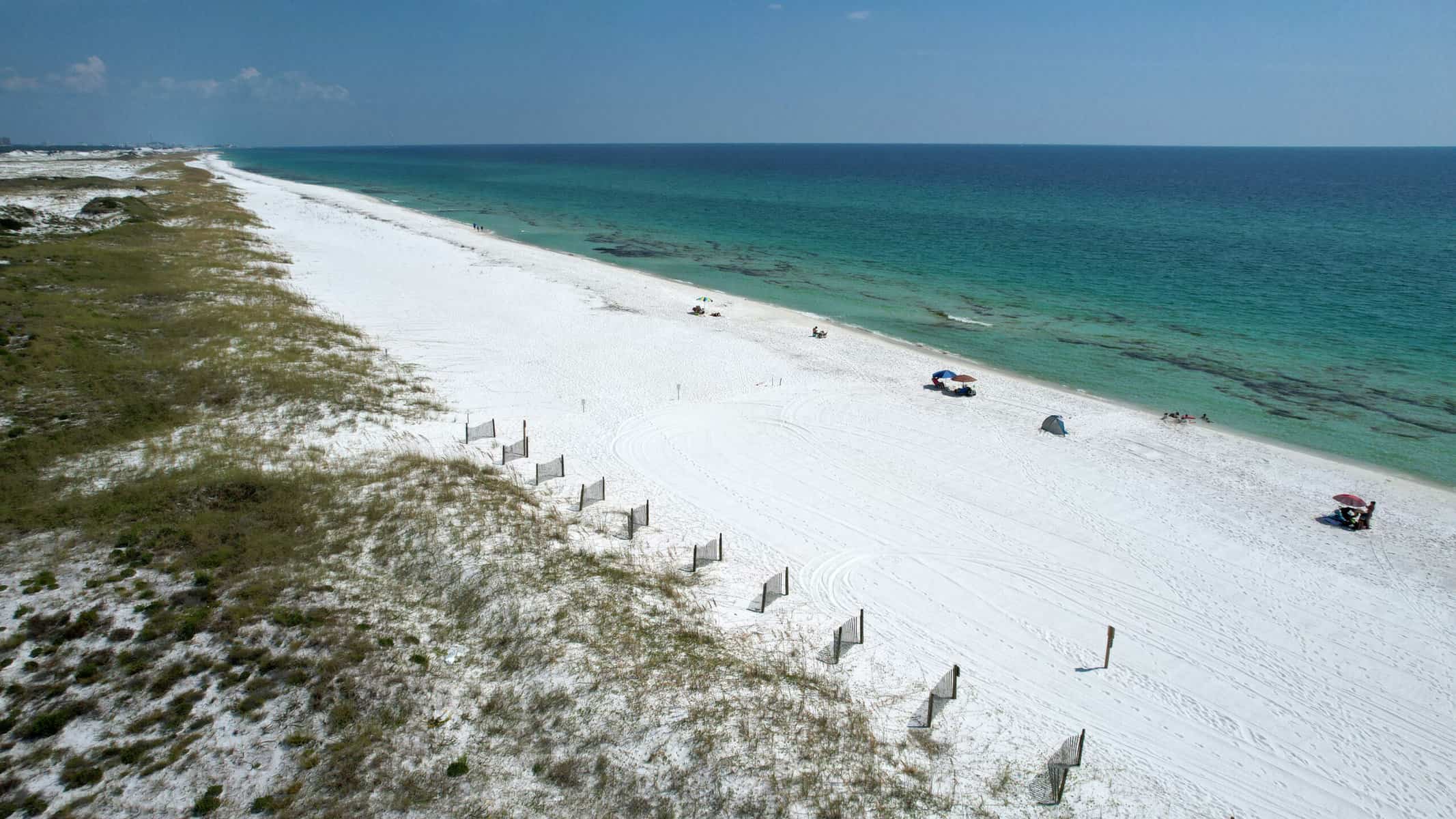 Aerial view of a sandy beach with clear turquoise water. The shoreline is lined with grass patches, and a few people are relaxing under small umbrellas. The sky is clear, adding to the serene and open atmosphere.