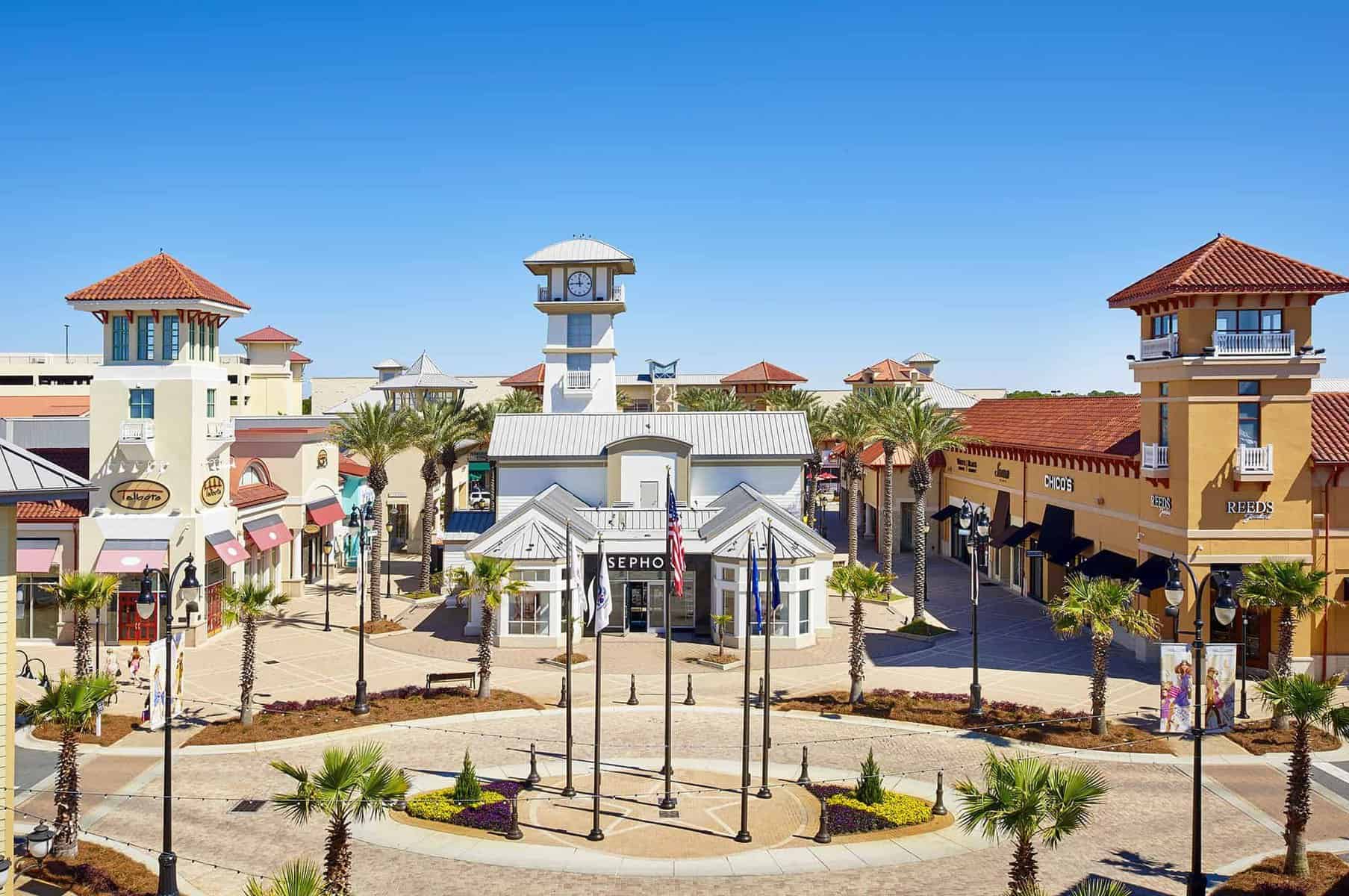 Outdoor shopping center with a round plaza, palm trees, and light-pink buildings featuring shops like Sephora and Pandora. The scene includes a central white tower with a clock and a clear blue sky overhead.