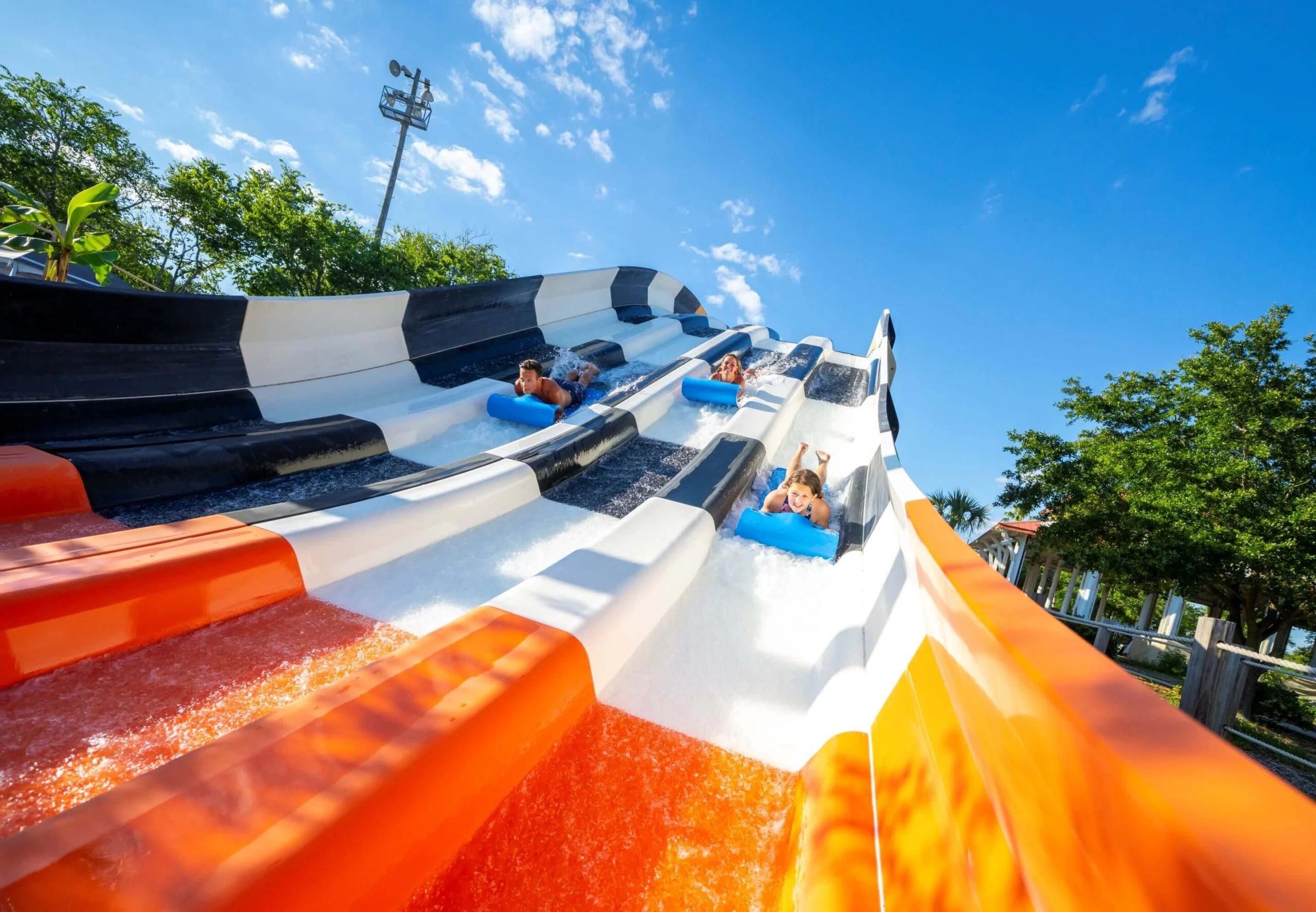 People are sliding down a large, multi-lane water slide on blue mats at a water park. The slide features black, white, and orange lanes, and the sky is clear with some clouds and surrounding greenery.