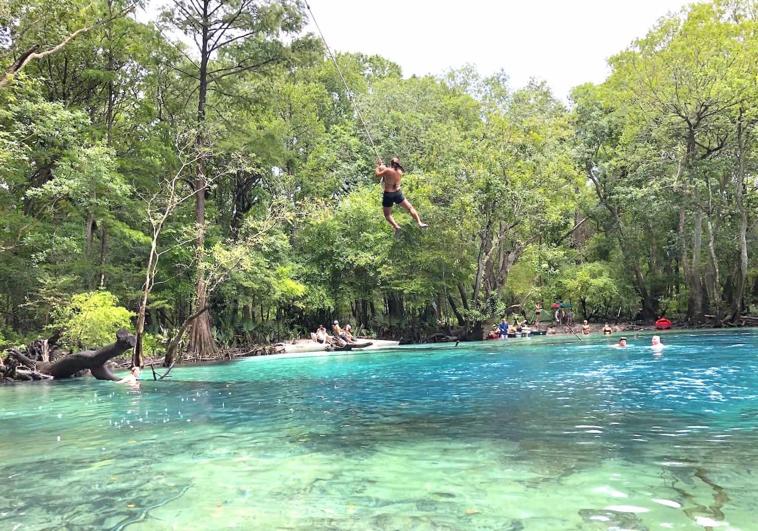 Person swinging on a rope over a clear blue forest lake. People swim and relax along the water's edge surrounded by lush green trees. Bright, sunny day.