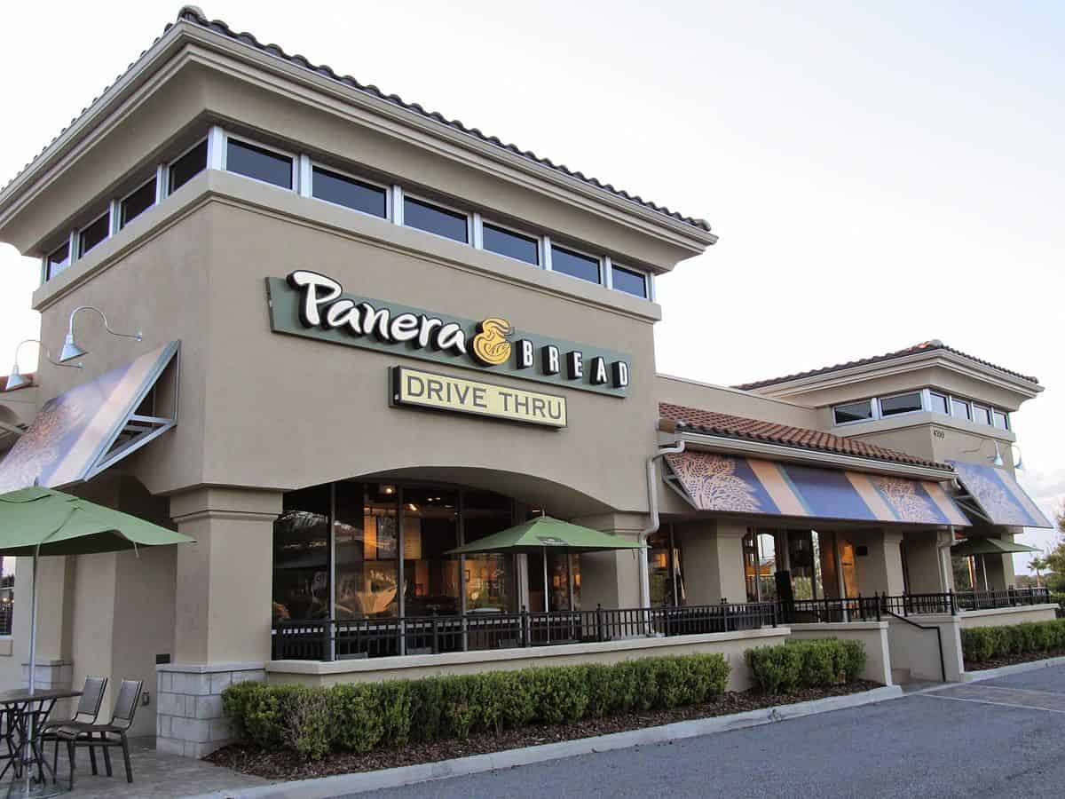 A Panera Bread restaurant with a drive-thru sign, featuring beige exterior walls, large windows, and green umbrellas over outdoor seating. The building has a tiled roof and landscaped greenery in front.