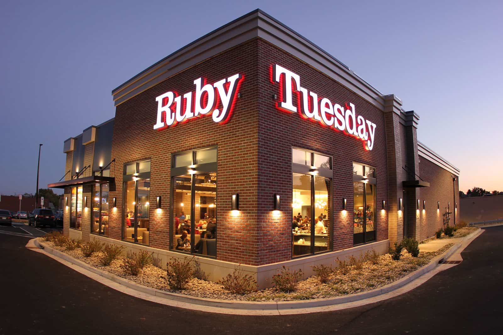 Exterior view of a Ruby Tuesday restaurant at dusk, featuring a brick building with large windows and prominent red and white signage. The parking lot is visible, and the warm interior lights create a welcoming atmosphere.