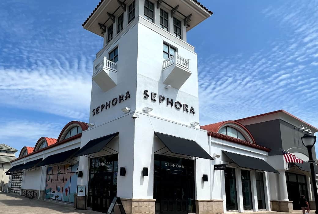 A large Sephora store with a prominent tower structure under a blue, cloud-streaked sky. The building has white walls, black awnings, and the Sephora logo in bold letters above the entrance.