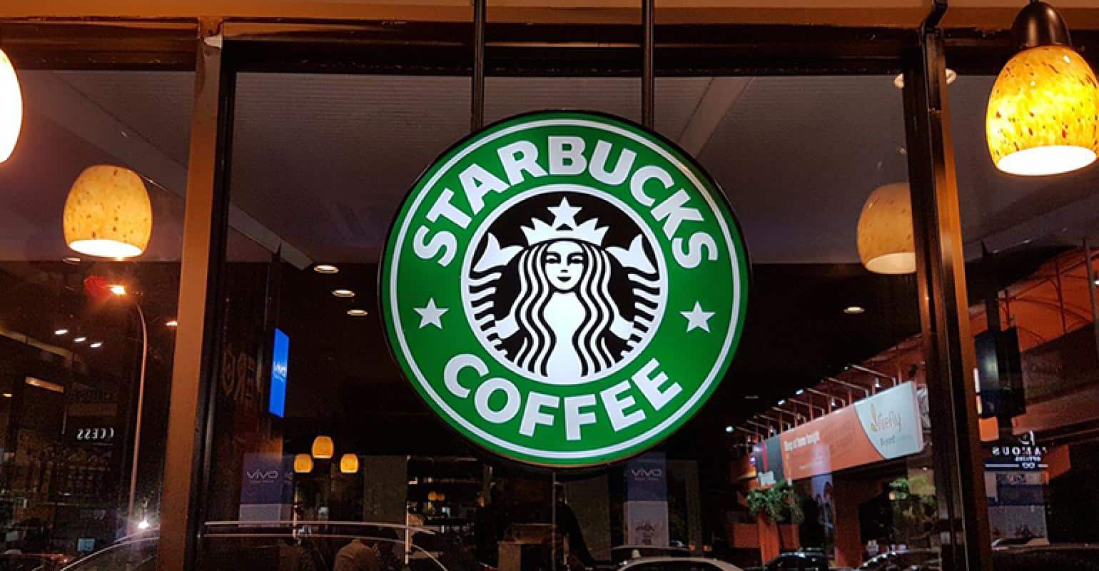 A Starbucks Coffee sign with the brand's green and white logo featuring a twin-tailed mermaid is displayed in a storefront window. Warmly lit hanging lamps are visible inside, and city streetlights and buildings are seen outside.