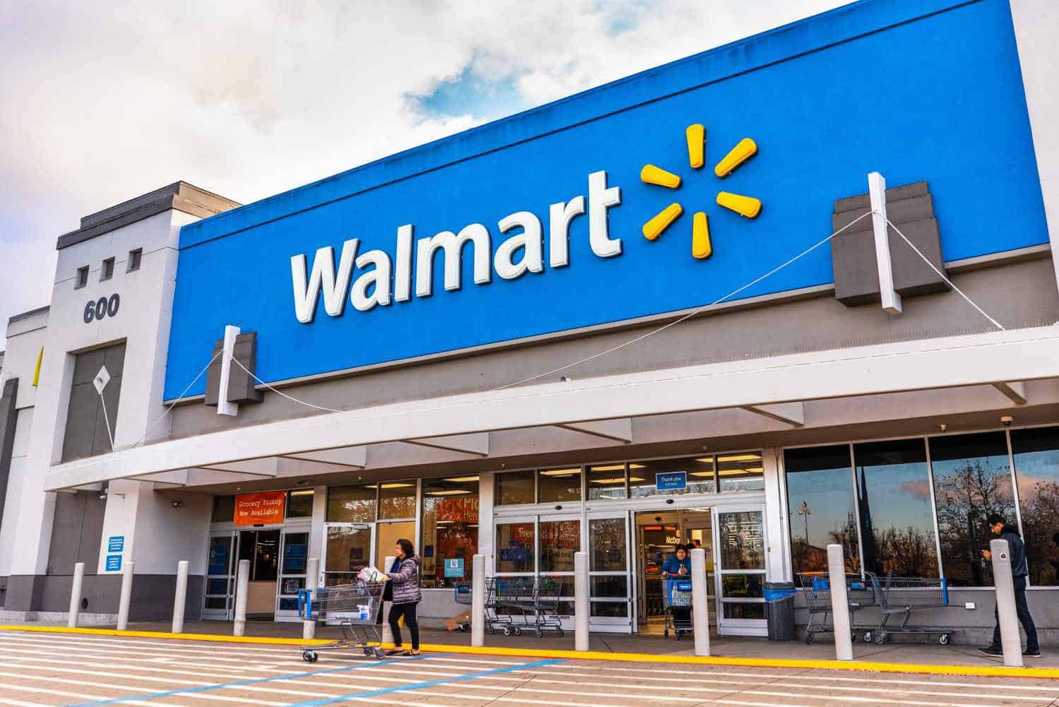 A Walmart storefront with a blue sign and yellow logo above the entrance. A person pushes a shopping cart toward the entrance. The store's address number, 600, is displayed on the building.
