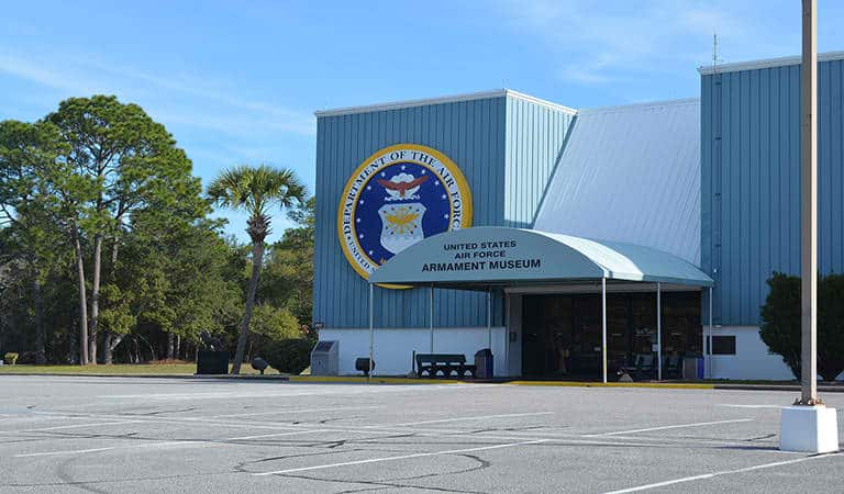 Front view of the United States Air Force Armament Museum. The building is light blue with a large Air Force emblem above the entrance. A clear sky and trees are in the background, and an empty parking lot is in the foreground.