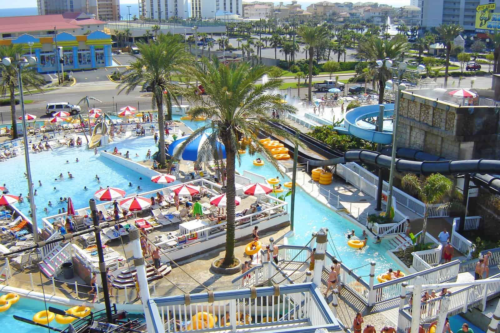 A lively water park with multiple pools, water slides, and a lazy river. People are enjoying the sun, swimming, and lounging under red and white umbrellas. Tall palm trees are scattered throughout the area, with buildings visible in the background.
