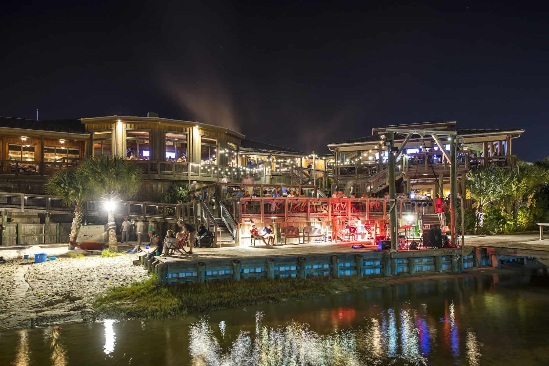 Nighttime scene of a waterfront restaurant with people dining on outdoor terraces. Soft lighting illuminates the wooden structure and surrounding palm trees. Reflections shimmer on the water below, creating a warm, inviting atmosphere.