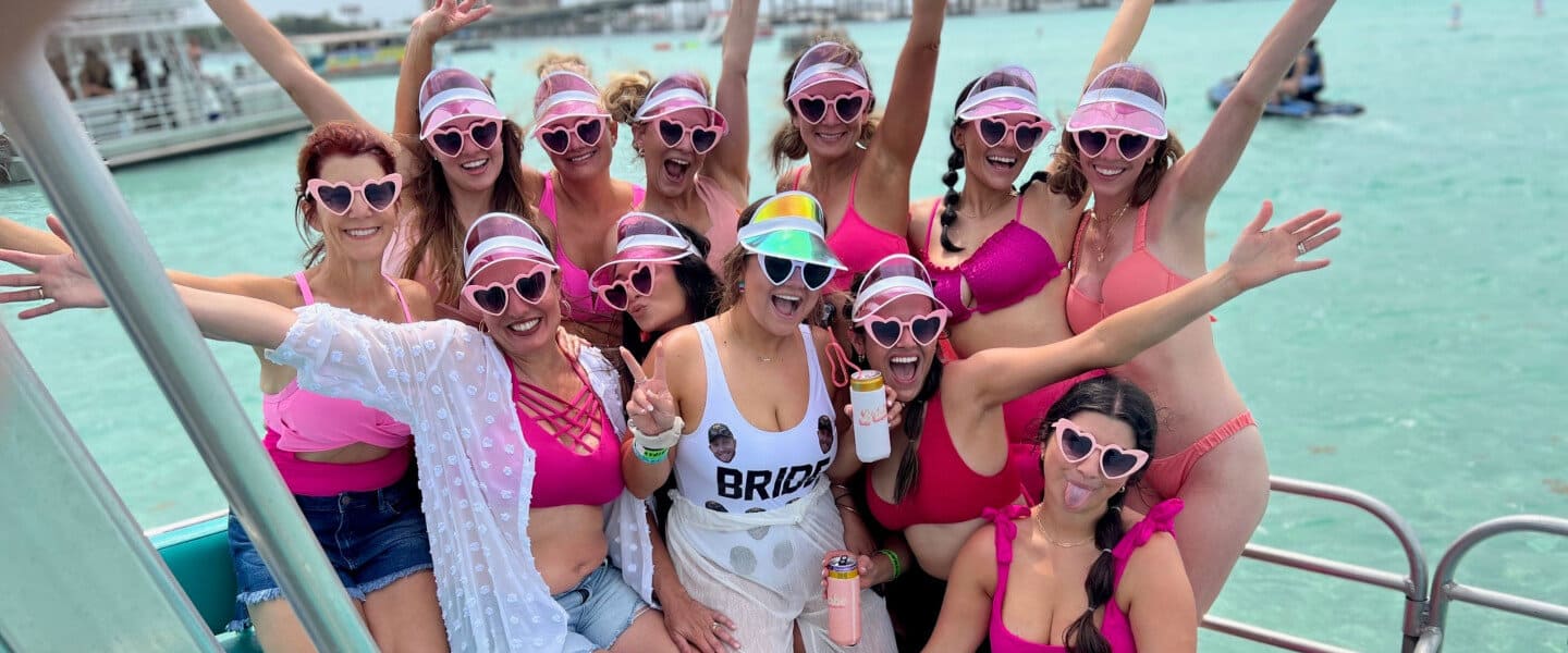 A group of women enjoying a boat trip, wearing pink bikinis and heart-shaped sunglasses. One woman in the center wears a 