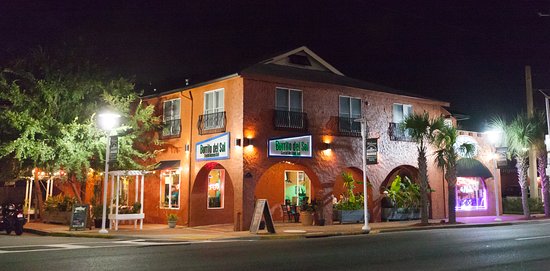 A two-story brick building at night with lit signage. The ground floor features large windows and plants near the entrance. Streetlights illuminate the exterior, and a neon sign is visible in a nearby window.