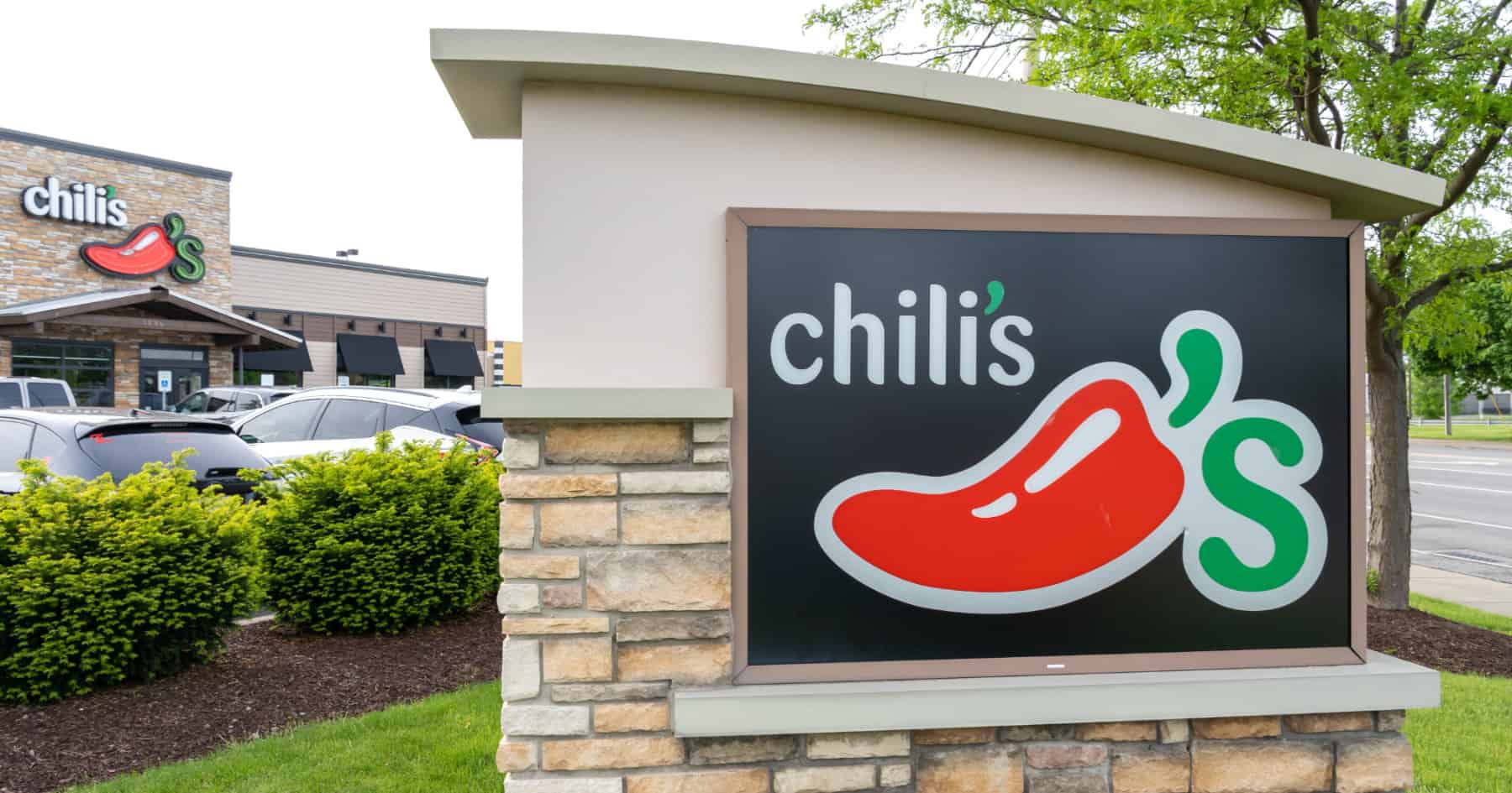 Sign for Chili's restaurant featuring a red chili pepper logo and text. The sign is mounted on a stone base with greenery around. The restaurant building is visible in the background, along with parked cars.