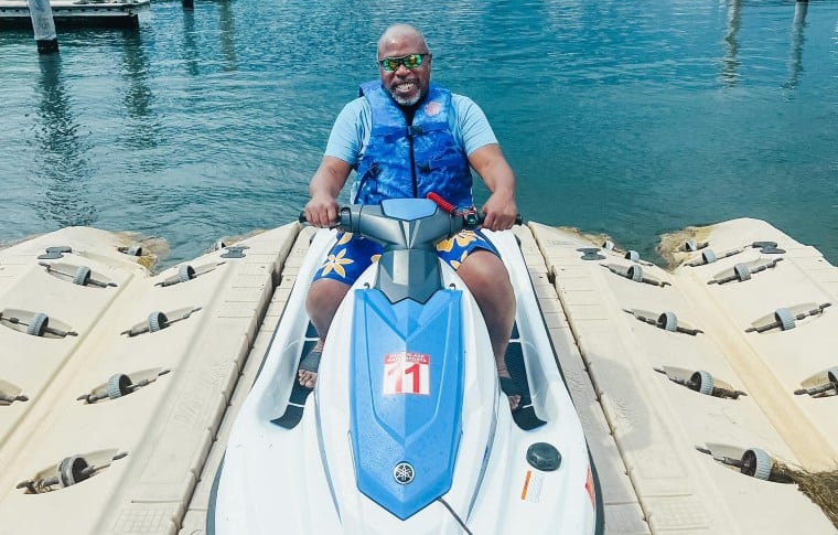 A man smiling while sitting on a stationary jet ski at a dock. He's wearing sunglasses, a blue life vest, and colorful shorts. The water is calm, and there are boats and piers in the background.