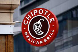 A circular red sign with the Chipotle Mexican Grill logo hangs on a building. The logo features a stylized chili pepper in white on a brown background, with