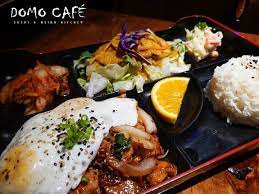 A meal tray from Domo Café featuring a sunny-side-up egg over seasoned meat, a side of salad with shredded vegetables, a serving of rice, a slice of orange, and pickled vegetables.