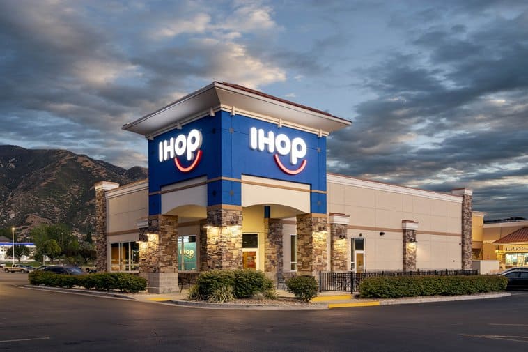 IHOP restaurant with a blue and white exterior and stone accents, located in a parking lot. The sky is cloudy, and mountains can be seen in the background. The IHOP logo is prominently displayed on the building.