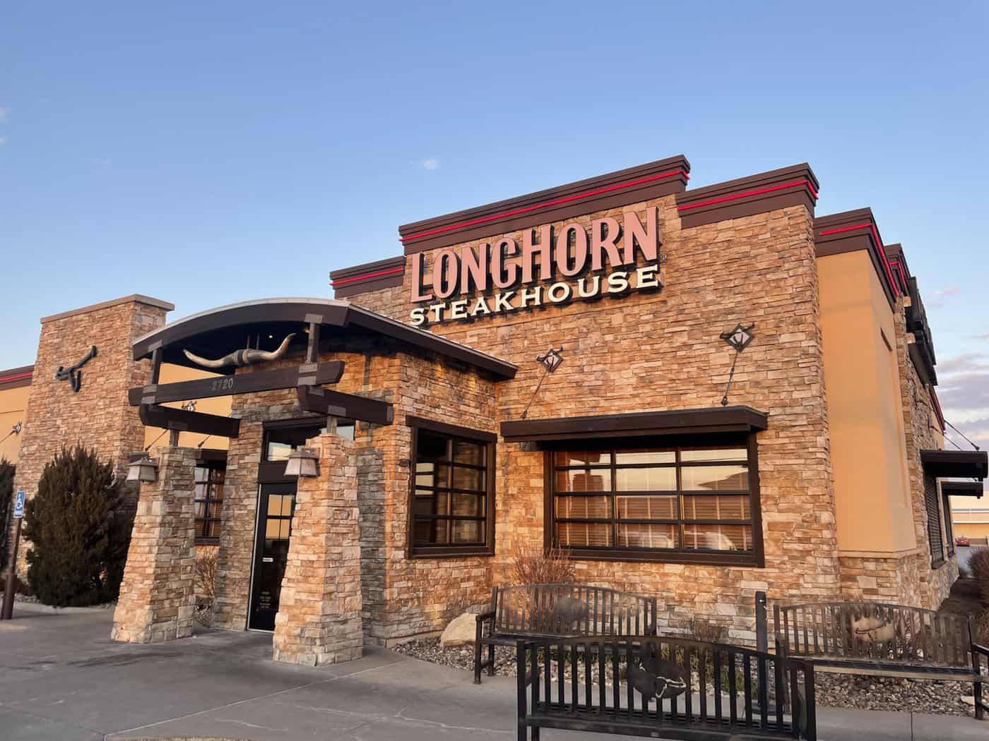 Exterior of a LongHorn Steakhouse with stone and wood accents. The building features western-themed decor, including a bull horn sign above the entrance. A bench is in front of the restaurant. The sky is clear and blue.