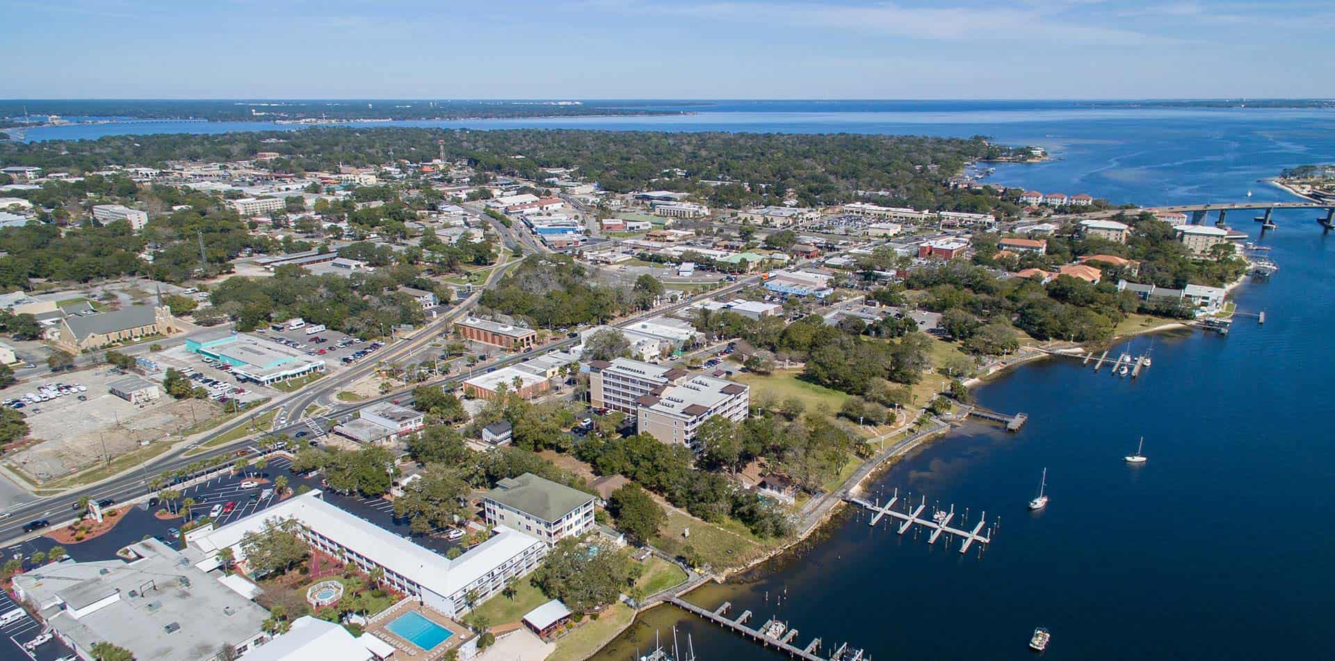 Aerial view of a coastal city with buildings, trees, and roads. A marina with boats is visible along the shoreline, and a bridge extends over the water. The horizon shows a wide expanse of blue water under a clear sky.
