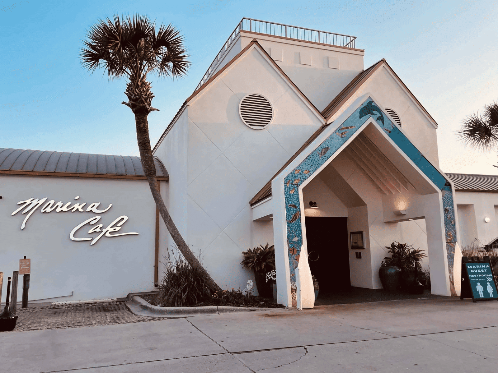 The image shows the exterior of Marina Café, featuring a tall triangular entrance with decorative blue accents. Two palm trees stand nearby, and the restaurant's name is displayed in stylish script on the left. The sky is clear at dusk.