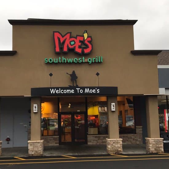Exterior of a Moe's Southwest Grill restaurant with a brown facade. The sign above the entrance reads “Moe's Southwest Grill” with a logo. Below, a sign says “Welcome To Moe’s.” The entrance has glass doors and stone pillars.