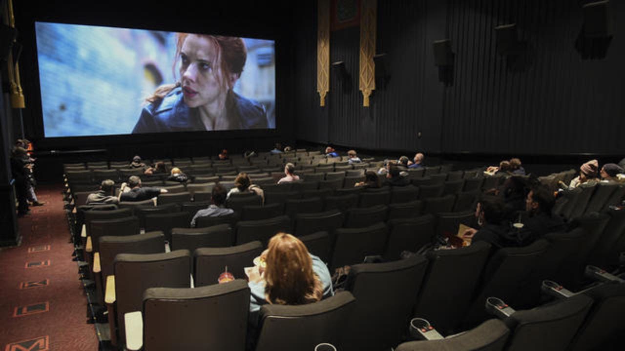 A dimly lit movie theater with patrons seated sparsely throughout the rows. A film is playing on the large screen, showing a scene with a person looking intently to the side. The theater's interior features dark walls and patterned carpet.