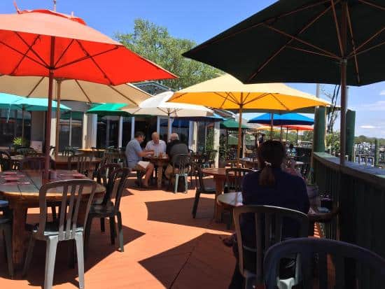 Outdoor cafe with colorful umbrellas providing shade over tables and chairs. People are seated at different tables, enjoying a sunny day. Trees and buildings are visible in the background.