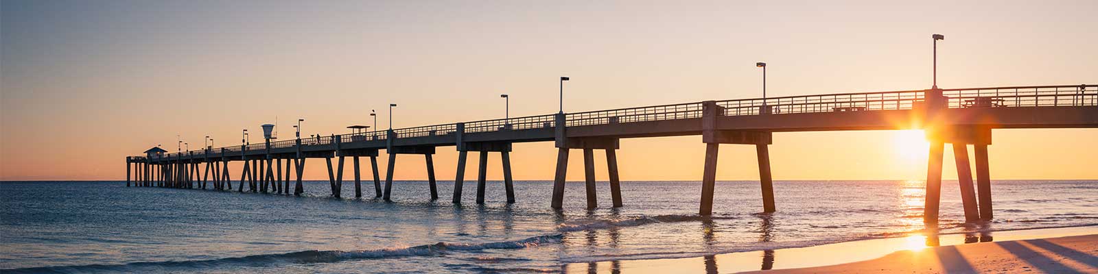A long pier stretches over the ocean at sunset. The sky is a gradient of warm colors, and the sun is partially visible on the horizon. Gentle waves lap against the shore, and the scene exudes a calm, serene atmosphere.