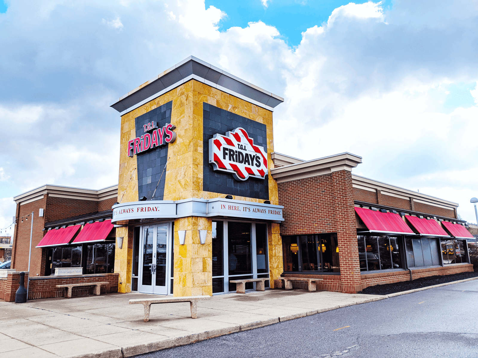 A TGI Fridays restaurant with a prominent sign on the corner. The building has a mix of yellow stone and red brick, with red awnings over the windows. A blue sky with scattered clouds is visible above.