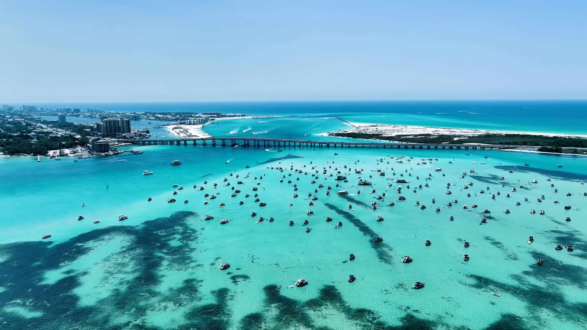 Aerial view of a turquoise bay dotted with numerous boats, a bridge stretching across the water, and a city skyline in the background. Sandy areas can be seen along the coast under a clear blue sky.