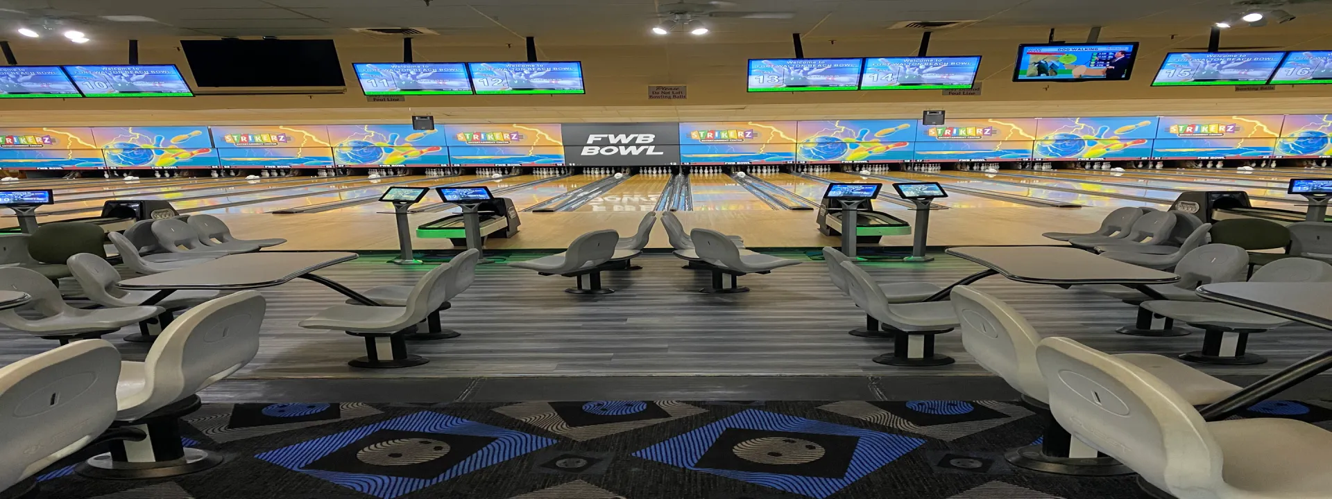 Interior of a bowling alley with several lanes. Monitors display colorful graphics above the lanes. Empty seating areas with chairs and small tables are seen in the foreground. The flooring has a geometric design.